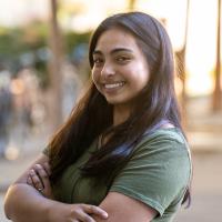 A young woman with long dark hair smiles confidently at the camera, standing outdoors with arms crossed. She is wearing a green T-shirt, and the background is softly blurred, with trees and sunlight visible.