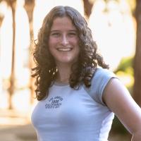 A young woman with curly hair and a friendly expression stands outdoors, smiling broadly at the camera. She is wearing a white and gray T-shirt with 'Los Angeles California Athletic Dept.' printed on it, with sunlight filtering through trees in the background.
