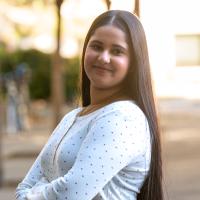 A young woman with long, straight dark hair and a gentle smile poses with her arms crossed, looking at the camera. She is wearing a white long-sleeve shirt with a small pattern, and the background shows blurred trees and a sunny outdoor setting.