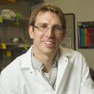A smiling man wearing glasses, a white lab coat, and a plaid shirt, seated in a laboratory setting with scientific equipment and colorful sticky notes in the background.