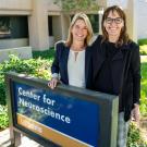 Two women standing in front of the Center for Neuroscience building next to the blue and gold Center for Neuroscience sign