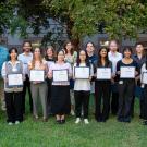 A group of sixteen individuals standing outdoors, each holding a certificate, with trees and a building in the background. They are dressed in business-casual attire, smiling for the photo, and are arranged in a single row on the grass.