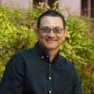 Portrait of a smiling person with short hair and glasses wearing a black button-up shirt with white polka dots, standing in front of green foliage.