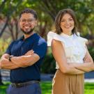 Two smiling coworkers standing side by side outdoors, arms crossed; man in navy, woman in white