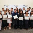 A group of UC Davis students pose together indoors, smiling and holding certificates during an award ceremony hosted by the College of Biological Sciences. A branded backdrop with the UC Davis logo is visible behind them.
