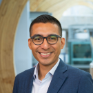 A smiling man wearing glasses and a navy blue suit jacket over a white dress shirt, standing in a modern building with curved wooden architecture in the background.