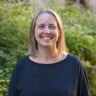 Person smiling outdoors in a black long-sleeve top, blond hair to shoulders, greenery and building behind.