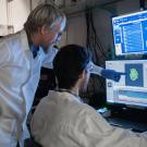 Two researchers in lab coats look at computer screens with one pointing to an image of cells on the monitor