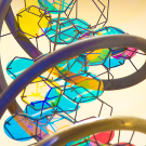 A close-up view of a colorful, abstract sculpture featuring intertwined metallic loops in dark and bronze tones. Within the loops are irregularly shaped panels resembling shards of stained glass in blue, yellow, and pink hues. Thin black lines connect the panels, creating an intricate web-like structure. 