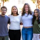 Four young adults stand side by side outdoors with their arms around each other, smiling warmly at the camera. The group consists of two men and two women, all casually dressed and appearing relaxed and friendly. Sunlight filters through trees in the background, casting a gentle glow on the group, who are standing on a paved path with greenery and a building partially visible behind them.