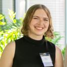 A smiling person with short, wavy light brown hair wearing a sleeveless black turtleneck top. They are standing indoors with green plants in the background and wearing a name badge that reads “Angelica Guerrero” with a ribbon labeled “Student Recipient.”