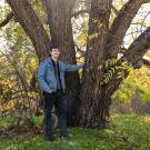 A person in a denim jacket leans casually against a large tree with multiple trunks, surrounded by grass and autumn foliage in a sunlit outdoor setting.