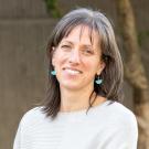 A woman with shoulder-length gray hair, wearing turquoise earrings and a light-colored sweater, smiles at the camera with a blurred outdoor background.