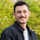 A smiling person with short dark hair, a mustache, and gold hoop earrings is outdoors in front of green foliage. They are wearing a black denim jacket over a white t-shirt. The background consists of lush green leaves, slightly blurred.