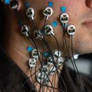 Close-up of a participant’s face and neck with multiple electrodes attached using blue medical tape. The electrodes are connected to thin black wires, leading to a monitoring device, highlighting a scientific study on muscle or nerve activity.