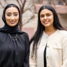 Two young women stand side by side outdoors, smiling at the camera. One wears a black hijab and a black blouse with gold necklaces, while the other has long black hair and wears a cream-colored blazer over a black top. The background features a blurred building and trees.