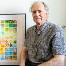 Richard Michelmore is seated to the left of a framed piece of art made of a grid of multicolored squares.