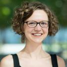 Portrait of a smiling individual with curly brown hair, wearing glasses, a black sleeveless top, and large earrings, set against a blurred outdoor background with greenery and soft lighting.
