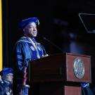 A university official in blue and gold academic regalia delivers a speech at a graduation ceremony, standing behind a podium with a university seal, while graduates listen in the background.