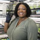 A smiling woman wearing gloves holds a small crab in a laboratory filled with terrariums and plant enclosures.
